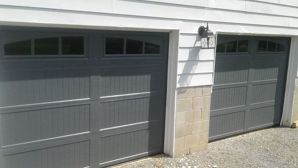 Two gray garage doors with windows, set against a white wall and gravel driveway.
