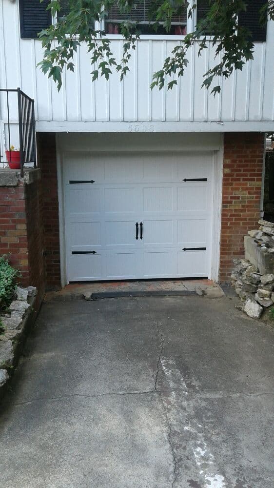 White garage door with black handles, brick and stone driveway, surrounded by greenery.