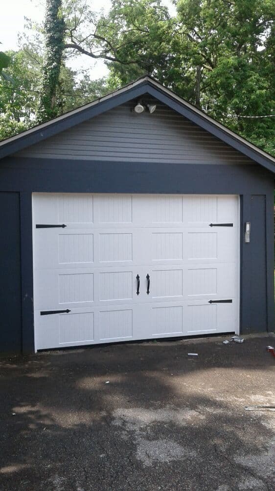 White garage door with decorative handles on a dark gray garage in a green, wooded area.