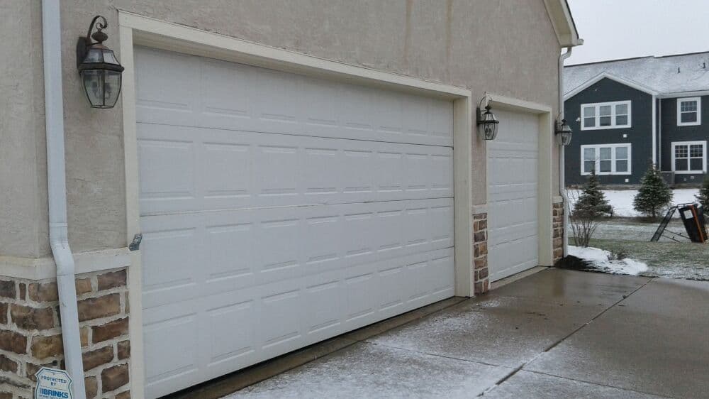 Two white garage doors with stone accents, snowy driveway, and suburban home background.