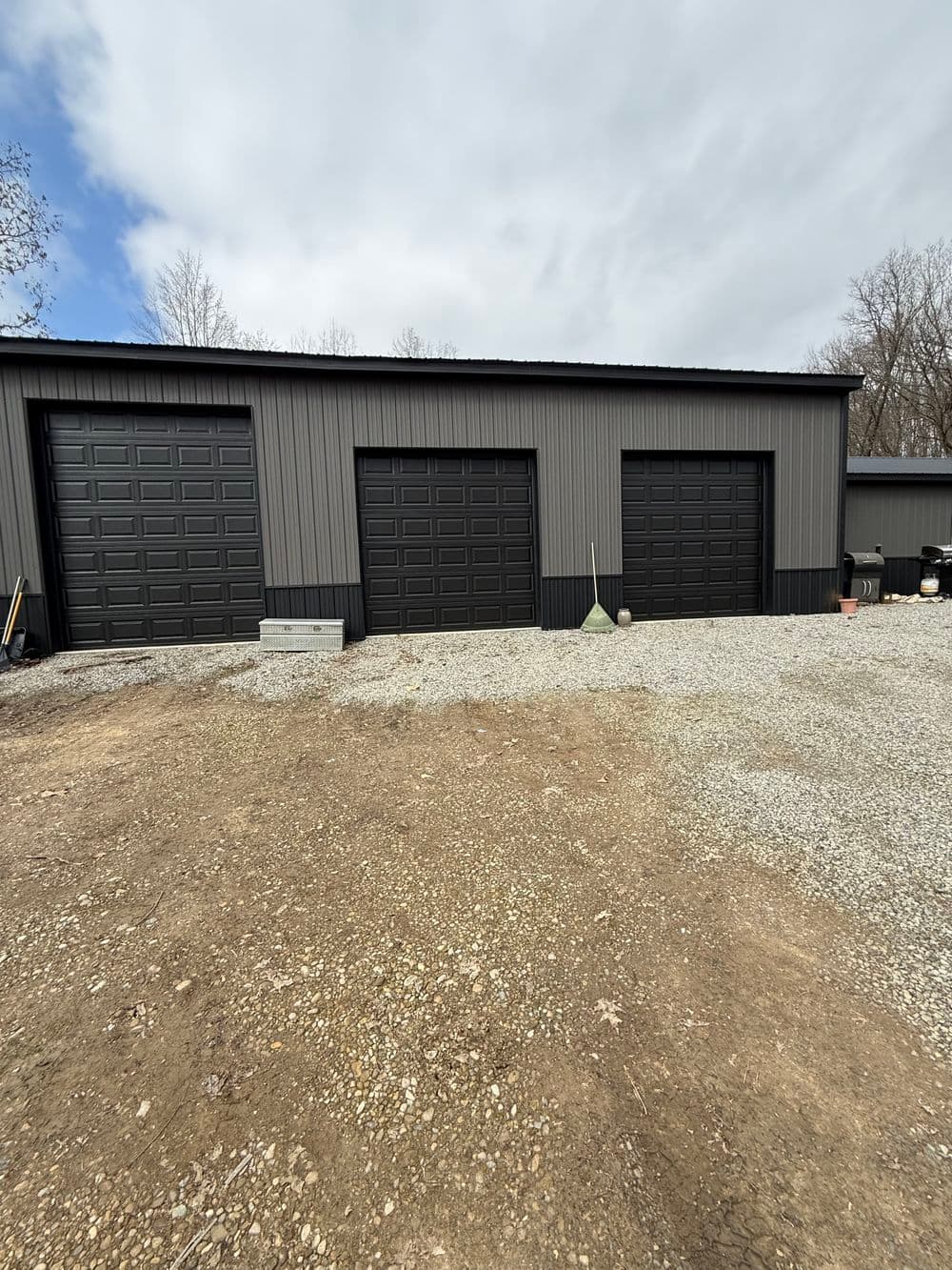 Modern metal garage structure with three black doors on gravel driveway. Cloudy sky above.