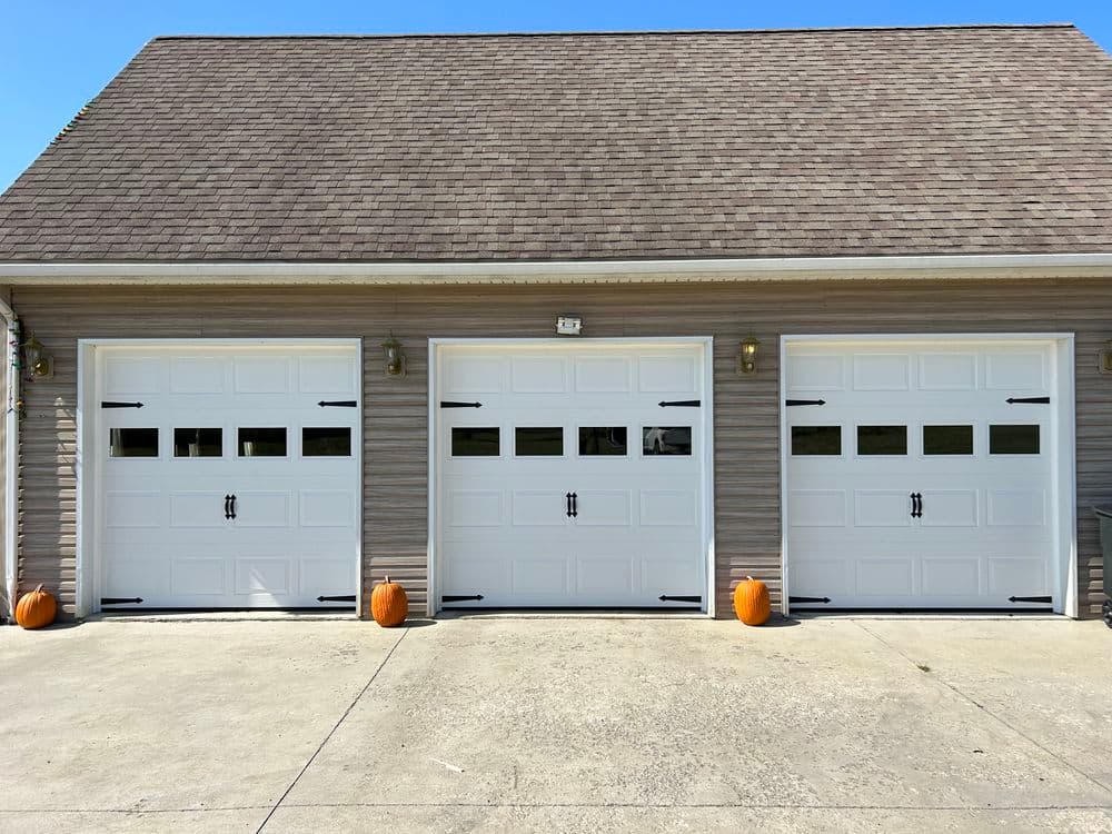 Three white garage doors with windows and pumpkins in front, set against a suburban home.