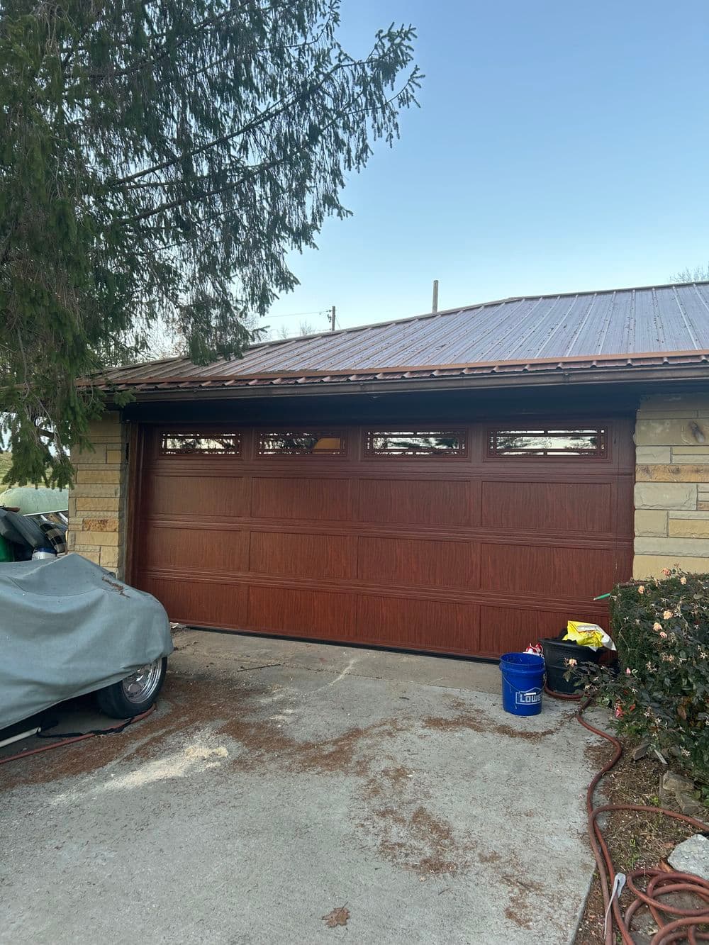 Brown garage door with windows and a metal roof, surrounded by greenery and a parked car.