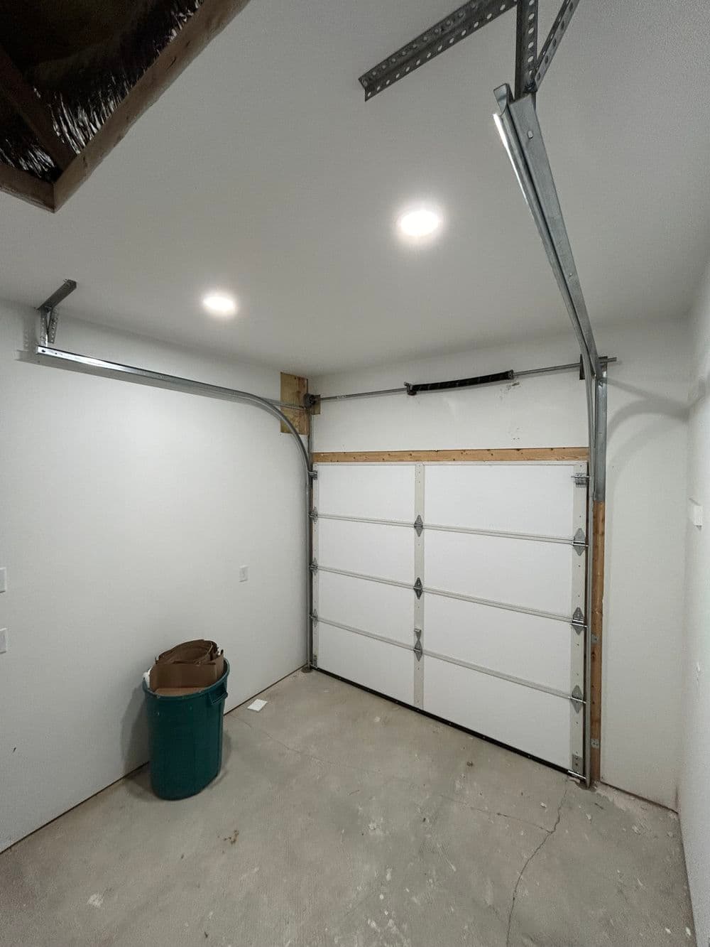 Empty garage interior with white walls, overhead lighting, and green trash bin.
