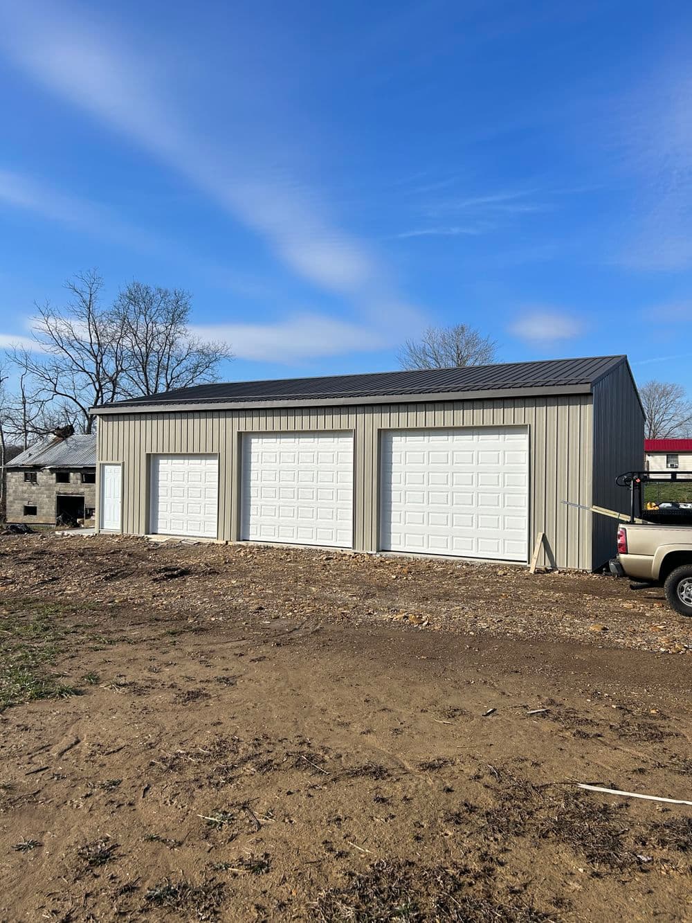 Three-door metal garage building on a gravel lot with truck in foreground and blue sky.